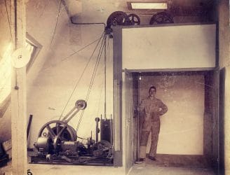Eine alte Fotografie zeigt einen Aufzugsführer in einer Aufzugskabine ohne Türen