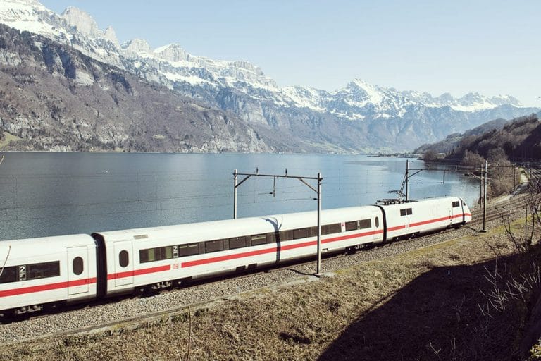 ICE-Zug fährt entlang eines Sees mit Bergen im Hintergrund
